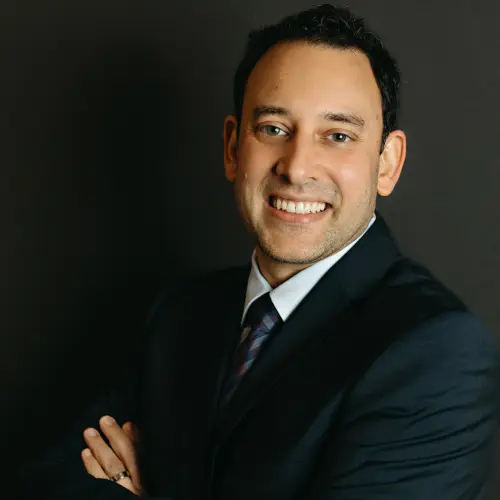 Dr. Neal Raval, smiling, in a dark suit, arms folded.