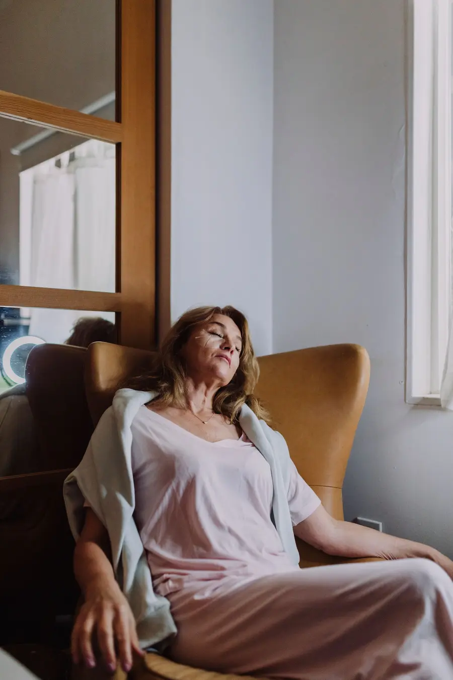 A woman resting in an armchair by a bright window, eyes closed.